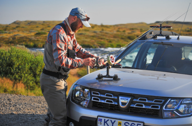 Man on a fishing trip in Iceland preparing his rods on the hood of a Dacia.